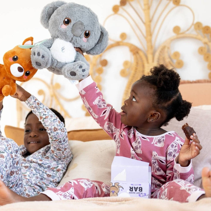 Children playing with plush toys on bed while eating a Sprout Organic snack bar.